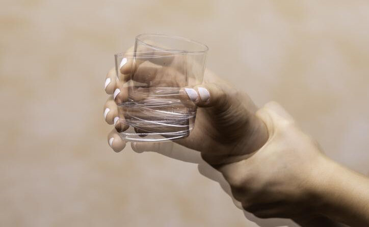 A shaking hand holding a glass and a second hand holding a hand for support; two glass figures against a peach background.