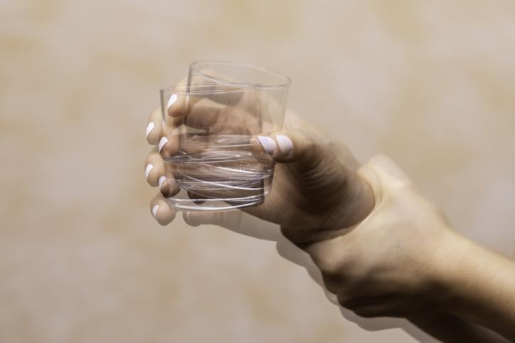 A shaking hand holding a glass and a second hand holding a hand for support; two glass figures against a peach background.